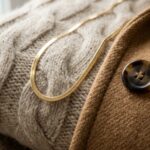 Ultra close-up macro documentary shot of luxury autumn wardrobe textures featuring folded cream cashmere knitwear, camel wool coat fabric, horn button, and delicate gold chain