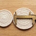 Four handmade cream cotton crochet coasters with brass caliper ruler measuring exact diameter on oak table