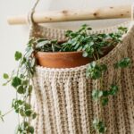 Close-up macro of handmade modern crochet wall pocket plant hanger mounted on off-white plaster wall holding four-inch terracotta pot with trailing leaves