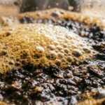 Ultra close-up macro of fresh coarse coffee grounds blooming with hot water and golden crema inside a glass French press