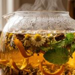 Ultra close-up macro eye-level shot of whole dried chamomile flowers, cinnamon bark, and mint steeping in warm amber tea liquid with rising steam