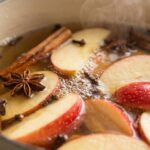 Ultra close-up macro of fresh red apples, cinnamon sticks and whole cloves simmering gently in cream Dutch oven