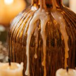 Ultra close-up macro of glowing ivory taper candles in vintage amber glass bottles with warm wax drips on rustic dinner table