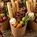 Extreme close-up macro of party individual charcuterie cups filled with artisan grissini breadsticks, halal beef salami ribbons, sharp cheddar cubes, olives, and rosemary