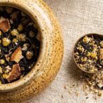 Ultra close-up macro of artisanal chamomile blossoms and loose black tea in ceramic canisters with antique brass measuring spoon
