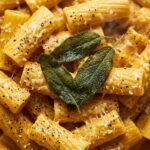 Ultra close-up macro texture of creamy garlic pumpkin pasta with ridged rigatoni, velvety orange sauce, grated parmesan, and crispy sage