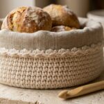 Close-up macro of handmade round crochet bread basket in double-stranded cream cotton with folded unbleached linen liner and warm dinner rolls on travertine workbench