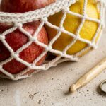 Close-up macro of handmade cream cotton diamond mesh crochet hanging fruit basket cradling red apples and yellow lemon on travertine workbench