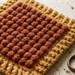 Macro close-up of handmade double-thick textured crochet hot pad showing raised bobble stitches and ribbed border on travertine stone