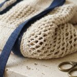 Macro close-up of handmade cotton crochet market tote bag showing interlocking open diamond mesh loops and dense single crochet base on travertine stone