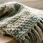 Ultra close-up macro photograph of handcrafted modern moss stitch crochet blanket in soft sage and cream super bulky yarn showing woven linen stitch texture