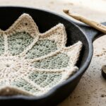 Close-up macro of handmade cream and sage starburst crochet pan protector resting inside a matte black seasoned cast iron skillet on travertine workbench