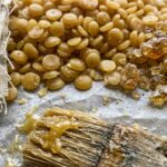 Natural golden beeswax pellets, amber pine rosin granules, and pre-cut patterned organic cotton fabric squares on parchment paper