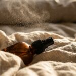 Ultra close-up macro of amber glass spray bottle misting aromatic droplets over neutral linen bedding