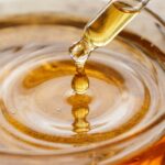 Ultra close-up macro of golden vanilla botanical body oil droplet rippling into glass dish