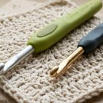 Close-up macro comparison of Clover Amour and Tulip Etimo 5.0 mm ergonomic crochet hooks resting on cream cotton swatch on travertine workbench