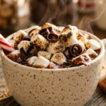 Extreme close-up macro of an artisan party hot cocoa mug filled with rich dark chocolate, piled high with toasted mini marshmallows, chocolate shavings, and peppermint stick