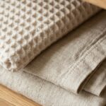 Ultra close-up macro texture of neatly folded waffle-knit cotton towels and neutral flax linen bedsheets on warm wooden shelf