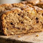 Ultra close-up macro food photography of freshly sliced moist cinnamon apple bread showing caramelized apple pieces, dark cinnamon swirl, and vanilla glaze on a rustic cutting board