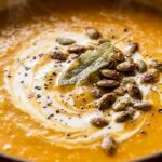 Ultra close-up macro food photography of velvety roasted butternut squash soup simmering with cream swirl and roasted pepitas