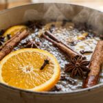 Ultra close-up macro eye-level shot of fresh orange slices, whole cinnamon sticks, star anise, and cloves gently simmering in a cream enameled cast iron pot with soft rising steam