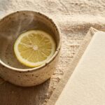 Ultra close-up macro of steaming ceramic mug of lemon water on linen runner beside open morning journal in dawn light