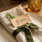 Extreme close-up macro of an elegant Thanksgiving place setting with cream ceramic plate, folded linen napkin tied with green velvet ribbon, fresh rosemary, and handwritten kraft name card