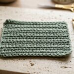 Close-up macro of handmade thermal single crochet fabric in sage green worsted cotton showing dense double-thick ridges and 4.5 mm wooden hook on travertine workbench